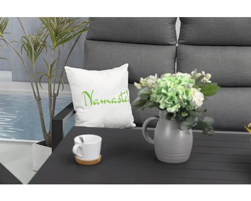 Dekorativní aranžmá s polštářem s nápisem Namaste, váza s květinami, šálek a rostlina na sedací soupravě