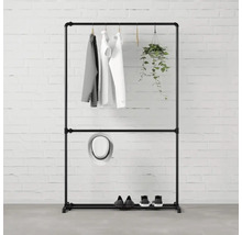 Černý stojan na šaty s oblečením, kloboukem, botami a závěsnou rostlinou před cihlovou zdí.