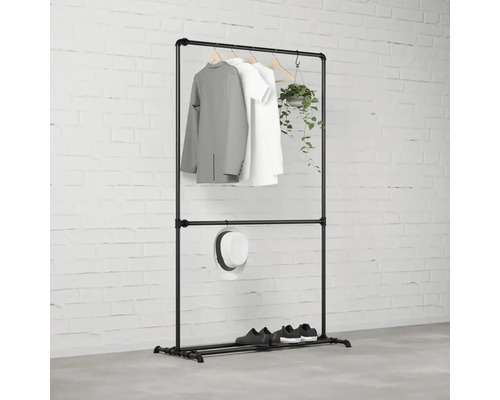 Černý věšák na šaty s oblečením, kloboukem, botami a rostlinou před cihlovou zdí