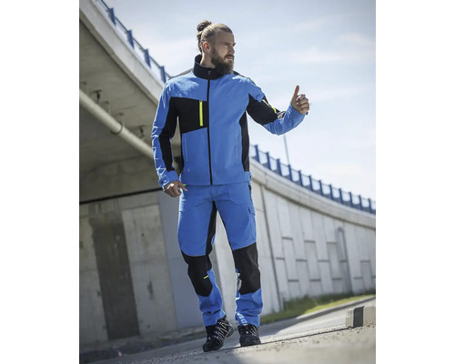 Muž v modré a černé pracovní bundě a pracovních kalhotách před betonovou zdí