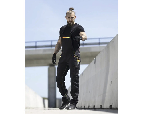 Muž v černém pracovním oblečení s lacláči před betonovou stěnou