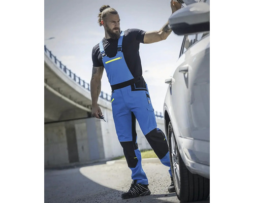 Muž v modrých pracovních kalhotách s laclem vedle auta.