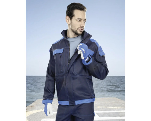Muž na sobě má tmavě modrou pracovní bundu s modrými akcenty a pracovní rukavice na pozadí moře.