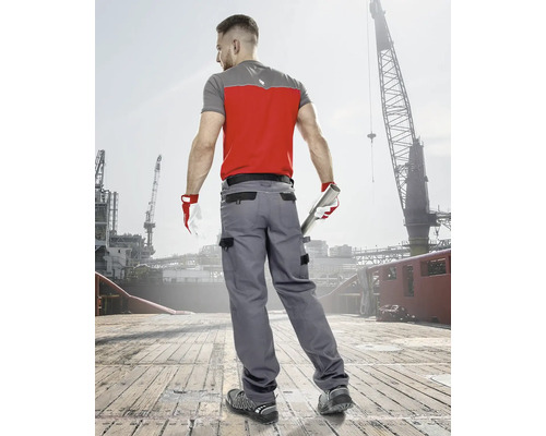 Muž v pracovním oděvu s tričkem, pracovními kalhotami, rukavicemi a bezpečnostní obuví před průmyslovou kulisou