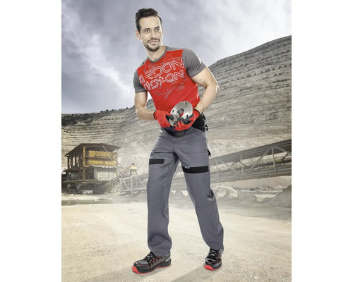 Muž v pracovním oděvu s pracovními kalhotami, tričkem a bezpečnostní obuví držící kovový díl