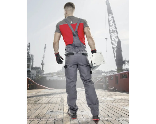 Muž v šedých pracovních kalhotách a červeném tričku drží kovovou desku před staveništěm.