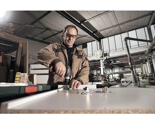 Muž montuje kování na kus nábytku vrtačkou v dílně.