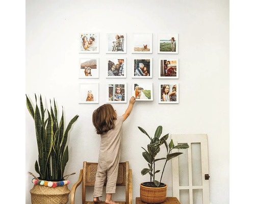 Dítě stojí na židli a dotýká se čtvercové fotokoláže na zdi, lemované pokojovými rostlinami.