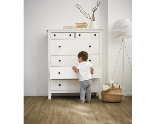 Dítě stojí před bílou komodou se zásuvkami a kulatými úchyty ve světlé místnosti.