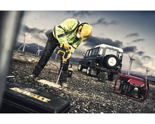 Stavební dělník vrtá vrtacím kladivem otvor do dřevěné desky. V pozadí je vidět terénní vozidlo a elektrocentrála.