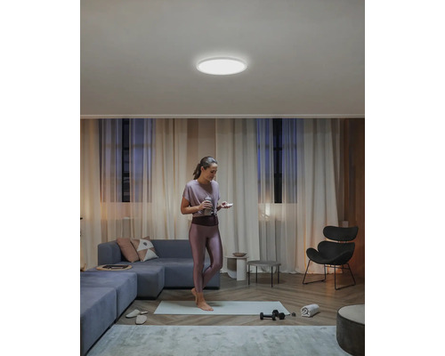 Žena stojí na podložce na jógu v obývacím pokoji s kulatým stropním svítidlem.