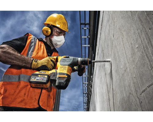 Stavební dělník vrtá vrtačkou otvor do betonové zdi. Dělník má ochrannou helmu, ochranné brýle, chrániče sluchu, obličejovou masku, výstražnou vestu a pracovní rukavice.