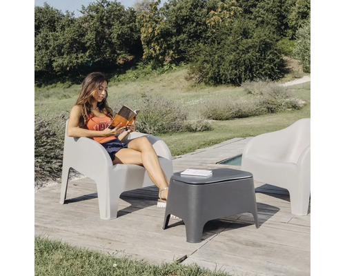 Venkovní scéna s ženou čtoucí knihu v šedém zahradním křesle a šedým zahradním stolkem vedle