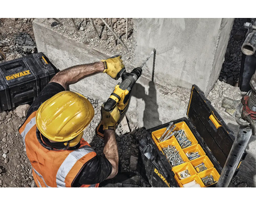 Stavební dělník vrtá vrtacím kladivem do betonové zdi, kufřík s nářadím a šrouby jsou připravené.