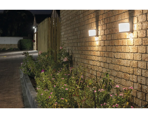 Dvě čtvercová venkovní světla na cihlové zdi vedle osázené plochy.