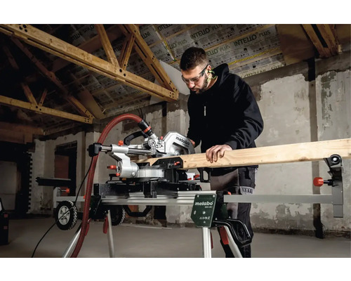 Muž s ochrannými brýlemi řeže dřevo pokosovou pilou Metabo na podstavci v hrubé stavbě.