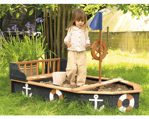 Dítě si hraje v pískovišti ve tvaru lodi s volantem a modrou plachtou