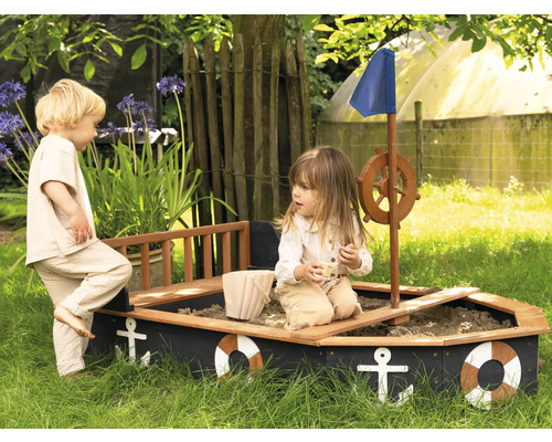 Dvě děti si hrají v pískovišti ve tvaru lodi na zahradě.