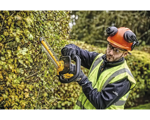 Osoba stříhá živý plot nůžkami na živý plot a má na sobě ochranný oděv.