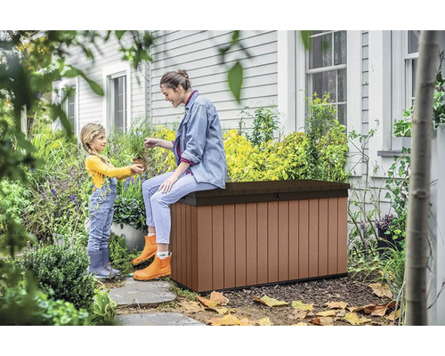 Zahradní scéna se ženou sedící na hnědé zahradní bedně a malým dítětem, které jí dává rostlinu.