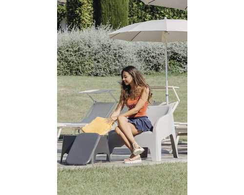 Venkovní scéna s ženou sedící na šedé zahradní židli s podnožkou, s žlutým ručníkem, slunečníkem a lehátky v pozadí.