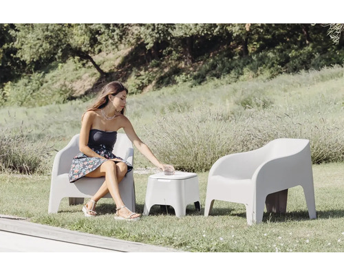 Scéna s ženou sedící na zahradním křesle, sahající po nápoji na zahradním stolku, vedle ní další zahradní křeslo na trávníku.