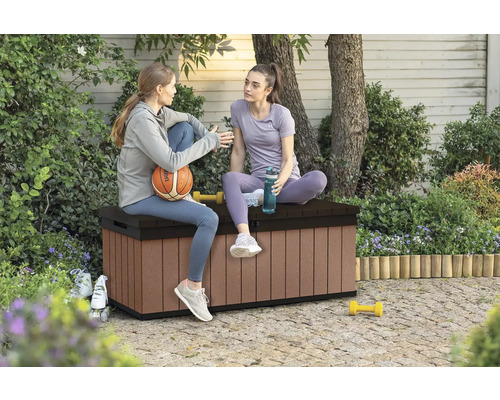 Dvě ženy sedí na hnědém plastovém úložném boxu na zahradě.