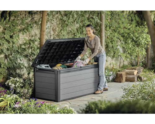 Zahradní scéna s ženou plnící šedý zahradní box venku. Box stojí na kamenné terase, obklopen rostlinami.