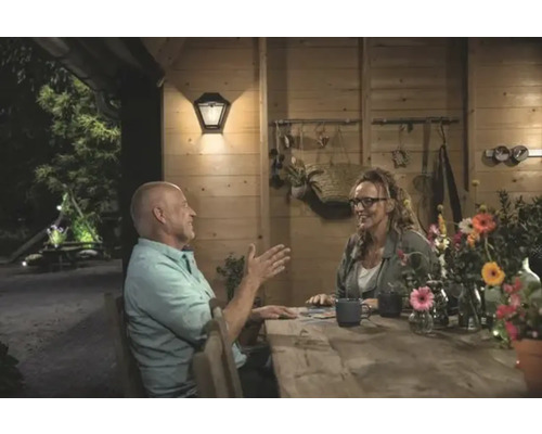 Pár sedí u dřevěného stolu v jasně osvětleném zahradním domku s venkovní nástěnnou lampou.