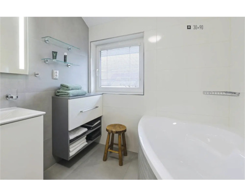 Světlá koupelna s vanou, umyvadlem, spodní skříňkou, ručníky, dřevěnou stoličkou a oknem se žaluzií