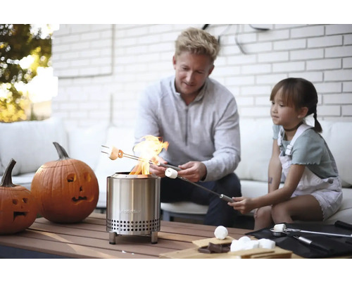 Otec a dcera opékají marshmallows nad venkovním ohništěm z nerezové oceli, zatímco na stole stojí vyřezávané dýně.