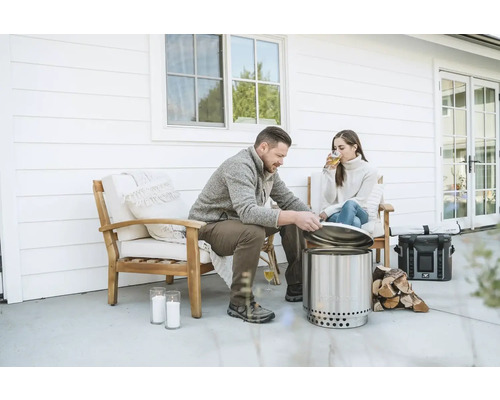 Scéna s mužem a ženou relaxujícími na terase, s nerezovým ohništěm bez kouře, palivovým dřevem a nápoji.
