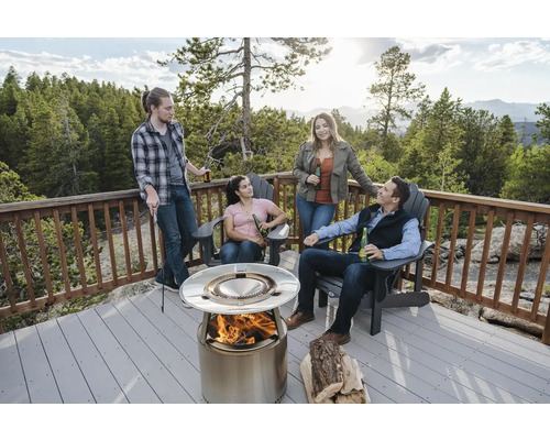 Scéna se čtyřmi lidmi shromážděnými kolem ocelového ohniště na terase se zábradlím, s kládami a židlemi