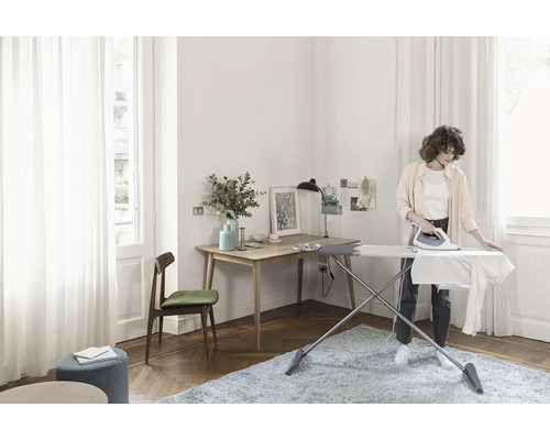 Světlá místnost s žehlicím prknem, žehličkou, psacím stolem a osobou při žehlení