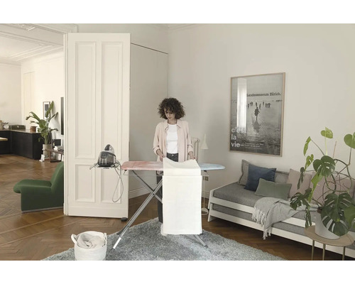 Žena žehlí prádlo na žehlicím prkně s napařovací žehličkou ve světlé místnosti.