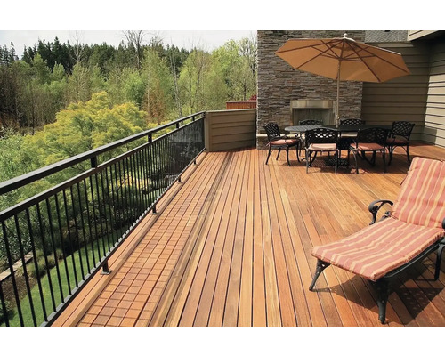 Prostorná dřevěná terasa se zábradlím, jídelní soupravou a lehátkem na zeleném pozadí.