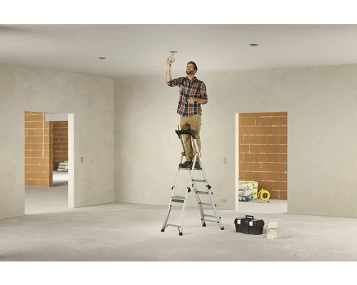 Muž montuje lampu na žebřík v nedokončené místnosti.