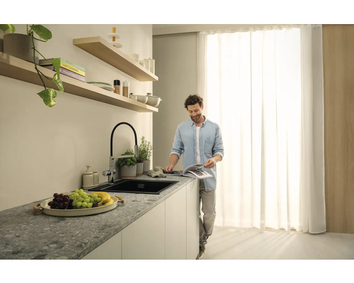 Světlý kuchyňský prostor s umyvadlem, vodovodní baterií a usmívajícím se mužem čtoucím knihu