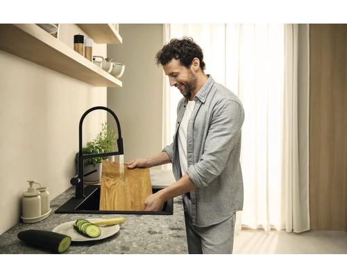 Muž myje dřevěné prkénko v černém kuchyňském dřezu pod černou baterií.