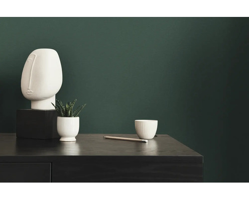 Dekorativní scéna se skulpturou, rostlinou, šálkem a tužkou na tmavém stole před zelenou stěnou