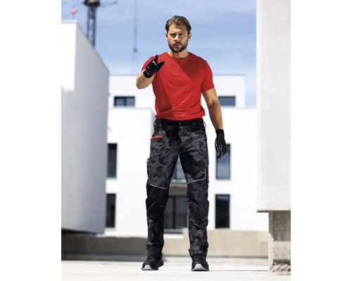 Muž v červeném tričku a maskáčových kalhotách s pracovními rukavicemi před budovami