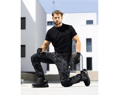 Muž klečí v pracovních kalhotách s maskovacím vzorem, černém tričku a rukavicích před budovou.