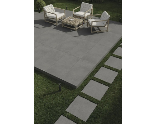 Terasa s šedou dlažbou, sedací souprava s dřevěným rámem a bílými polštáři a šlapáky v trávníku.