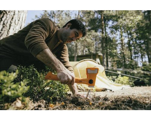 Muž používá kladivo k zatlučení kolíku do země, v pozadí je stan.