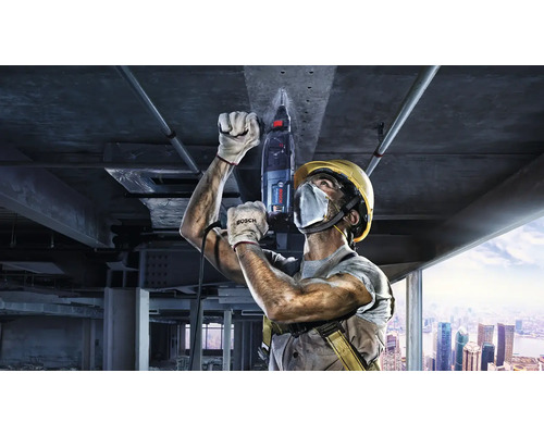 Stavební dělník vrtá do stropu vrtacím kladivem, v pozadí staveniště.