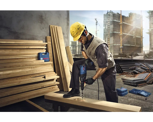 Stavební dělník vrtá vrtačkou otvor do dřevěné desky na staveništi.