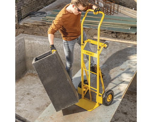 Osoba přepravuje šedou nádobu se žlutým rudlem na staveništi.