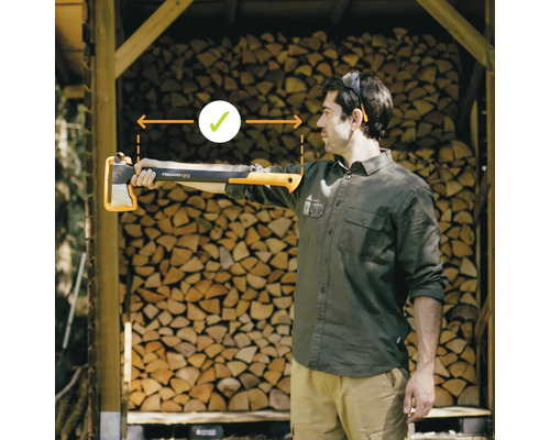 Muž drží štípací sekeru Fiskars před hromadou dřeva, aby demonstroval správnou délku pro práci