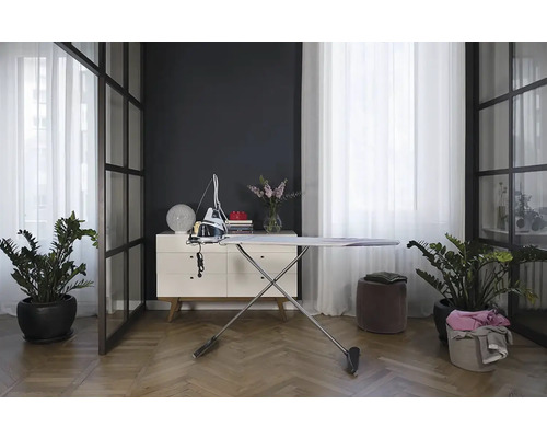 Žehlička a žehlicí prkno ve světlé, moderní místnosti s rostlinami a bílým příborníkem.