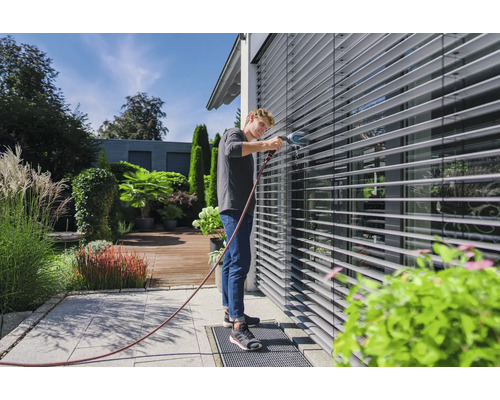 Muž čistí venkovní žaluzie kartáčem a vodní hadicí na zahradě.